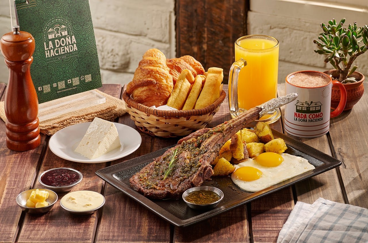 Tomahawk a la parrilla con chimichurri, papas y huevos fritos, junto a una canasta de pan, café y jugo, en La Doña Hacienda