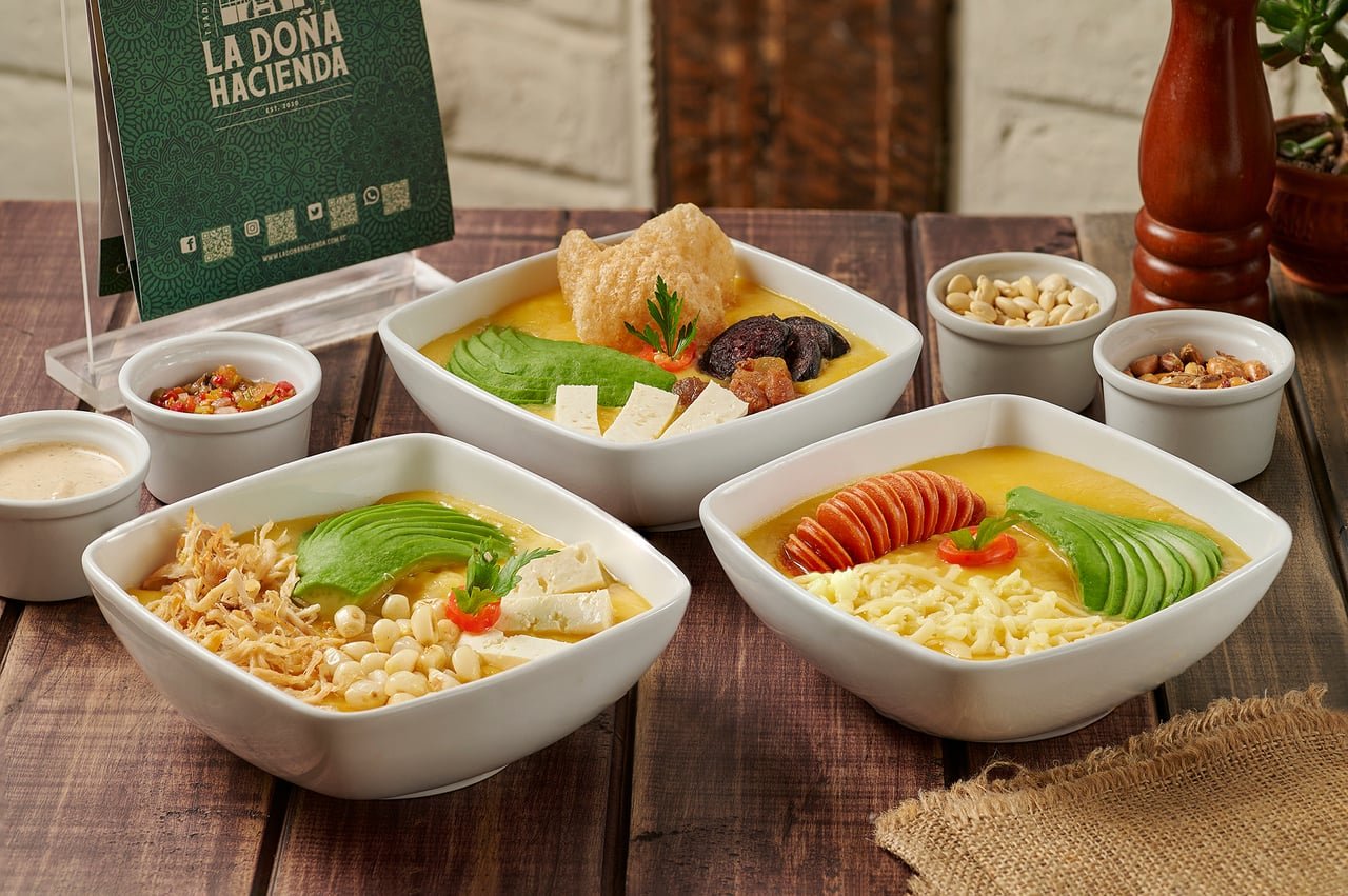 Tres tazones de sopa cremosa amarilla con aguacate, queso, chicharrón, chorizo y pollo deshilachado, servidos con maní y tostado en La Doña Hacienda