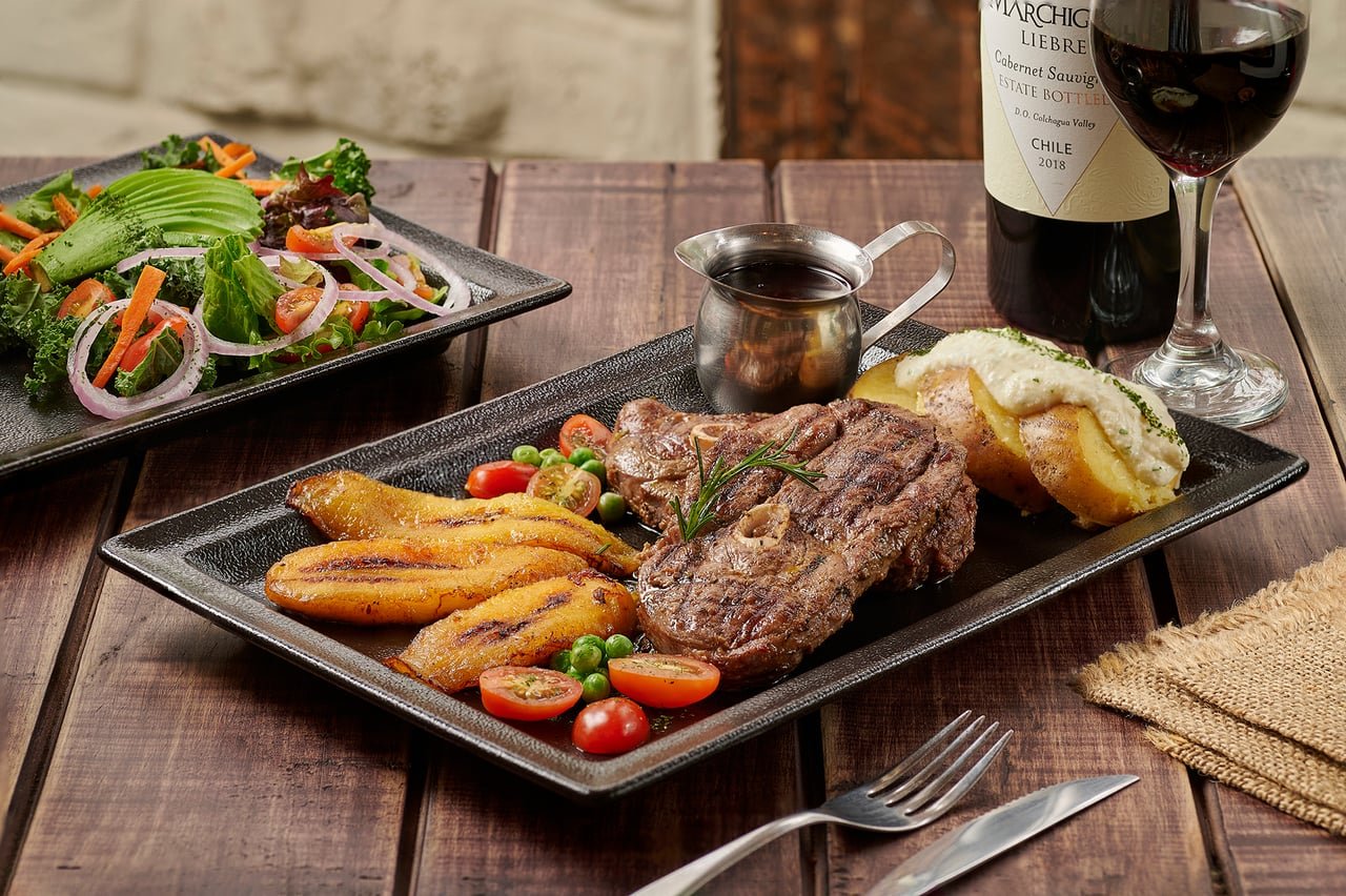 T-bone a la parrilla con maduro frito, papa al horno rellena y ensalada, servido con una copa de vino tinto, en La Doña Hacienda