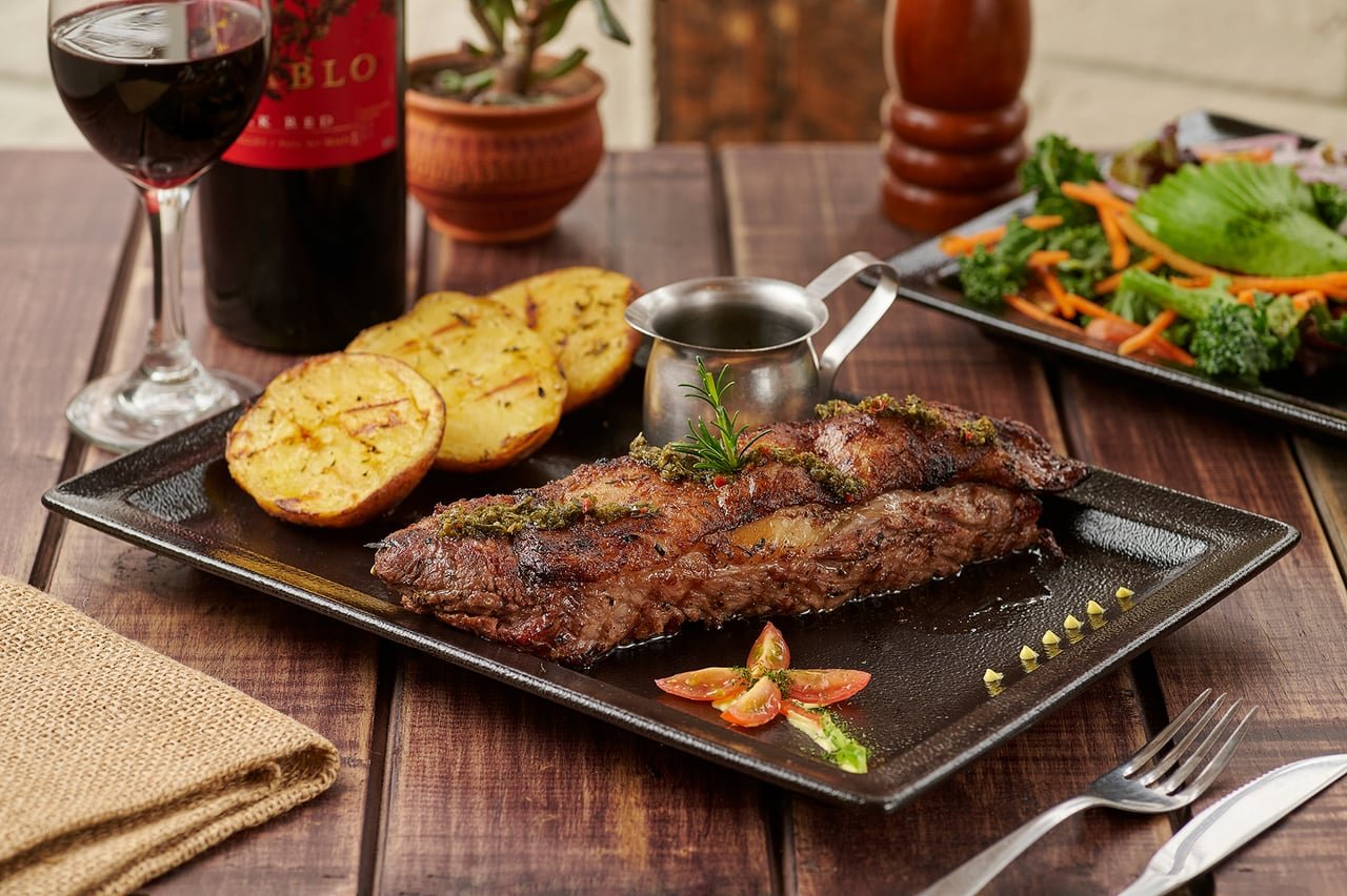 Churrasco a la parrilla con chimichurri, papas al horno y ensalada, servido con una copa de vino tinto, en La Doña Hacienda